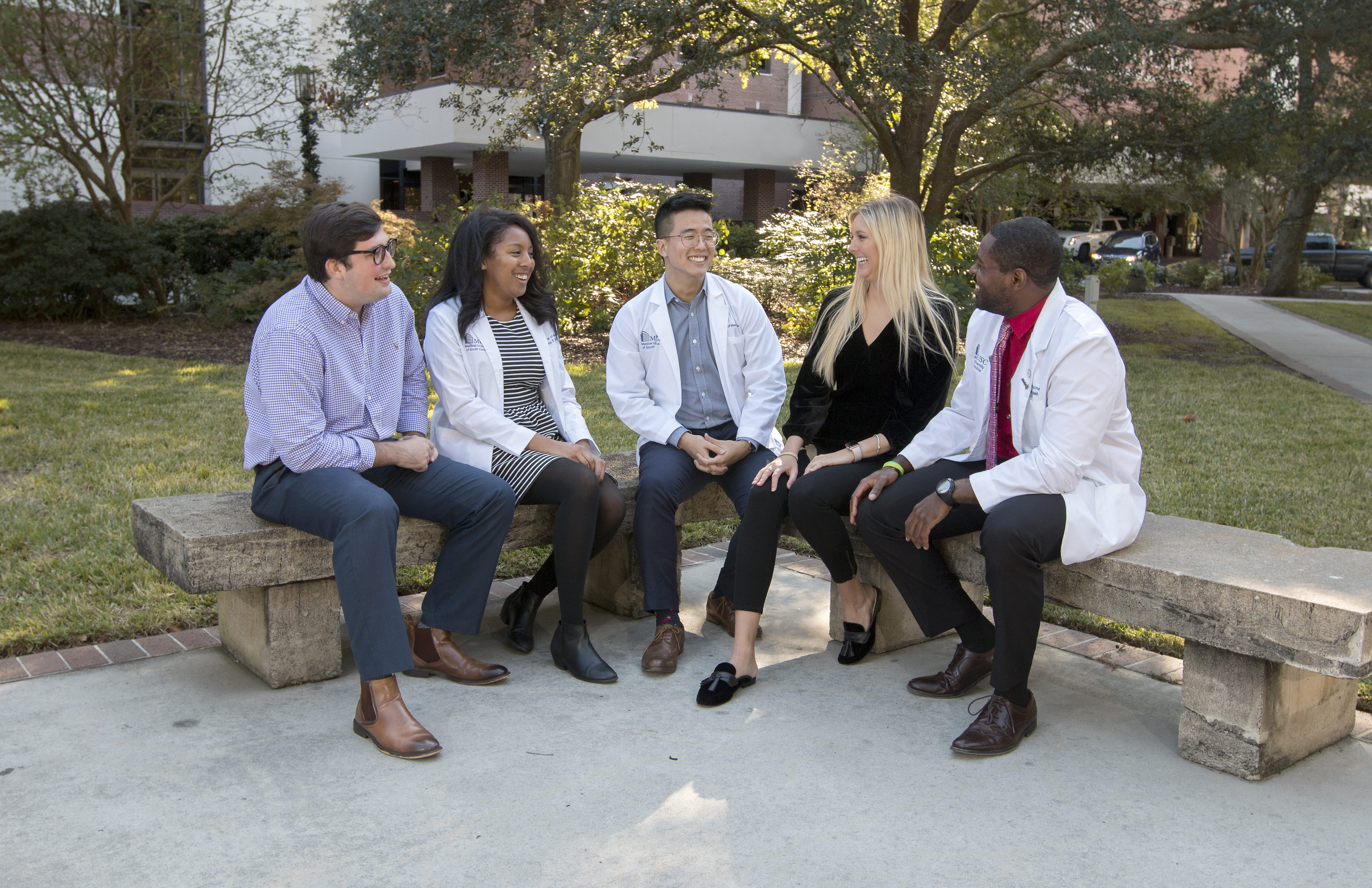 5 medical students wearing white coats sitting on bench on campus
