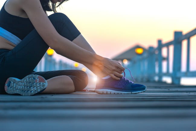 Image of a woman in exercise clothes, sitting on a pier while tying her sneakers as the sun rises in the background.