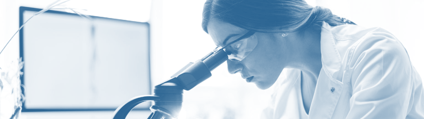 Photo of lab researcher with safety glasses on looking through microscope