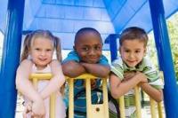 Three children smiling leaning over the top of a climbing structure on a playground