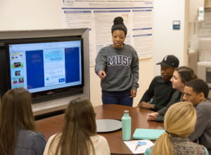 One MPH student presents information from a large monitor to six other students seated around the collaborative area
