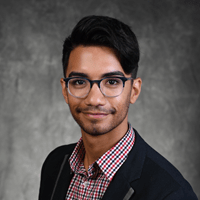 Christian Adames poses for a headshot against in a grey background wearing glasses and a plaid shirt.