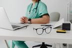 doctor sitting at desk with hands clasped and looking at laptop with glasses on the desk
