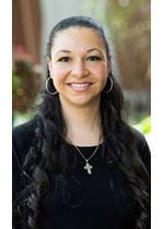 Ciara is wearing a long sleeve black shirt with a gold crucifix. She has long, black, curly hair and is smiling at the camera.