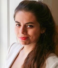 Marta is standing beside a window and is smiling. She has on a cream color blazer and her brown hair is pulled into a ponytail.
