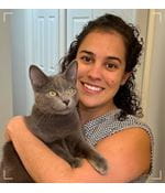 A woman is wearing a grey sleeveless blouse holding a grey cat that's looking off camera. The woman is smiling