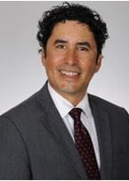 headshot of a man with curly black hair wearing a suit with a red polka dot tie