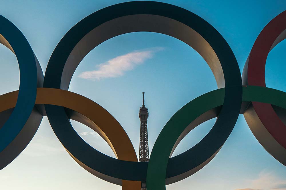 Olympic Rings are in foreground with the Eiffel Tower visible through them.