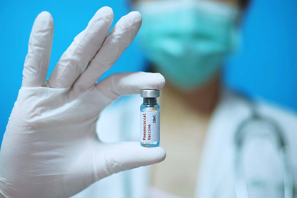 Masked woman wearing gloves holds vial of pneumonia vaccine.