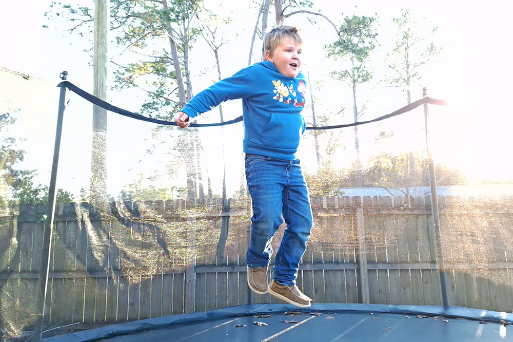 a young boy wearing a Sonic the Hedgehog sweatshirt jumps as high as he can on a backyard trampoline with the sun shining into the camera behind him