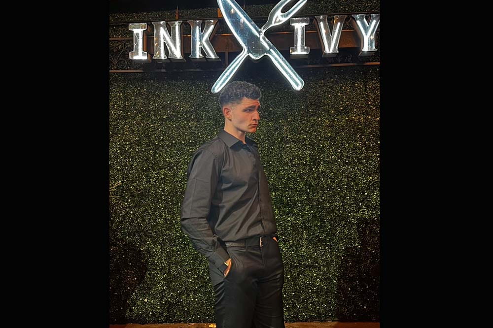 Young man wearing dress clothes stands in front of a sign that says Ink Ivy.