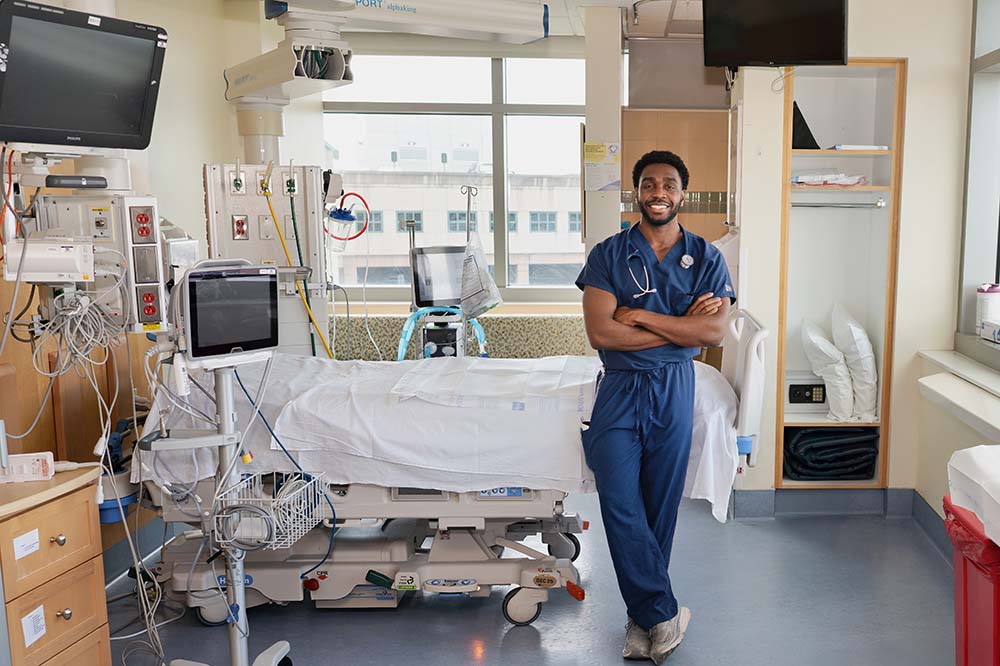 Man wearing hospital scrubs stands by a bed. There is medical equipment in the room.