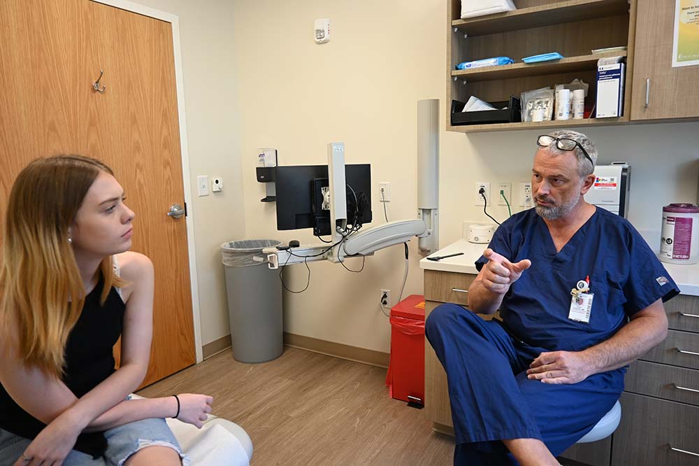A young woman with dark blonde hair wearing a black sleeveless top and jeans talks with a man wearing blue hospital scrubs in a clinic room.